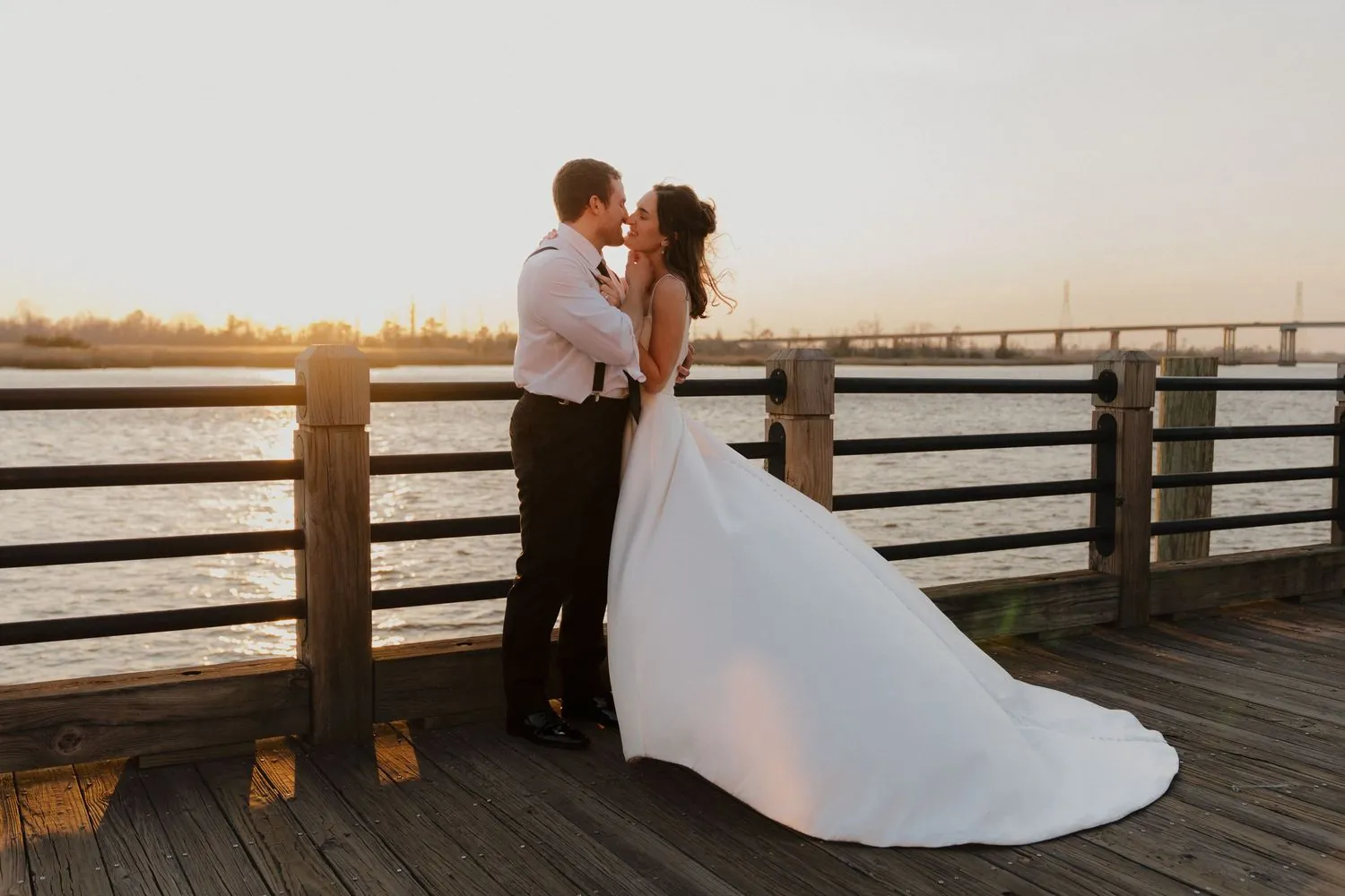 Bride and groom kissing at a waterfront railing at sunset, her gown spread across the dock